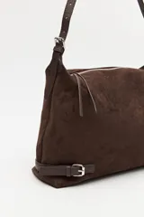 Cartera de gamuza negra con textura suave y acabado aterciopelado. Cuenta con una silueta amplia y flexible, cierre superior metálico, correa regulable y detalles laterales con hebillas.