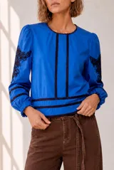 Blusa blanca de manga larga con detalles de bordados florales en tono grisáceo y bandas decorativas en el frente y mangas. Cuenta con cuello redondo y puños elásticos.
