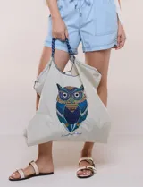 Bolso tote amplio color beige, con estampado frontal de un búho colorido. Cuenta con abertura superior ajustable mediante cordón y asas de cordada trenzada bicolor.