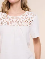 Blusa blanca de textura gofrada, de corte recto y semi holgado, con mangas cortas y escote redondo adornado con un canesú frontal en crochet calado.