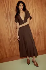 Vestido midi de color marrón, confeccionado en tejido de gasa con textura. Presenta un escote en pico con detalle de lazada frontal, mangas cortas abullonadas con terminación elástica y falda con detalle de godets que aportan volumen. Incluye forro interior a tono y cierre lateral con cremallera oculta.