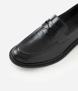 Mocasín Satinato de mujer confeccionado en cuero negro con acabado liso, puntera redondeada, costuras visibles, tira decorativa en la pala y suela con taco bajo.