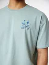 Camiseta blanca de algodón orgánico, con corte oversize, cuello redondo y manguita corta. Presenta un pequeño estampado rojo en el lateral superior izquierdo con la frase 'Go Out And Slide' y caracteres en japonés junto a una ilustración de dos personas cargando una tabla de surf.