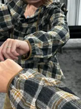 Camisa de manga larga con estampado a cuadros en tonos gris, negro y mostaza. Presenta volados en la parte superior delantera y en los hombros, con un diseño de corte holgado y lazos decorativos en el cuello.