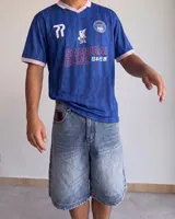 Camiseta de fútbol de manga corta en color azul con diseño estampado abstracto y cuello en V rosado con detalles blancos a juego con los puños. Presenta inscripciones y escudos deportivos en el pecho.