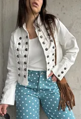Chaqueta corta de estilo militar en color blanco, con múltiples filas de botones metálicos grabados y detalles de cordones en el frente. Cuenta con cuello mao y mangas largas con botones en los puños.