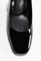 Zapato tipo bailarina o mary jane de charol negro, con punta fina y taco bajo de bloque. Posee una tira fina ajustable en el empeine con hebilla dorada.