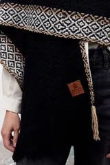 Ruana de tejido de punto en color negro, con diseño de guardas geométricas en tonos beige y blanco en los bordes y terminación con flecos.
