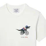 Remera de algodón blanca de corte clásico, con cuello redondo y manga corta. Presenta un pequeño estampado en el pecho que muestra al icónico cocodrilo de Lacoste vestido con ropa deportiva y montando una bicicleta, junto con los logotipos de Lacoste y Netflix.