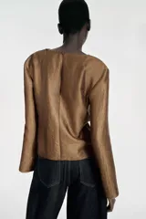 Blusa de manga larga con cuello fluido tipo drapeado y abertura en los puños. Presenta un acabado satinado con textura y cierre posterior con cremallera oculta.