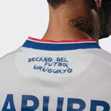 Camiseta de fútbol Umbro del Club Nacional de Football, modelo Home 2025, para hombre. Diseño blanco con sutil patrón en relieve, cuello redondo con ribetes azules y rojos, y detalles en las mangas. Incluye escudo del club, logo Umbro y sponsor Antel en el frente.