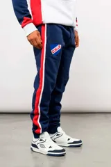 Pantalón deportivo tipo jogging de la marca Umbro, en color azul marino con bandas laterales en rojo y blanco. Cuenta con el escudo del Club Nacional de Football en el muslo derecho y el logo de Umbro en el izquierdo, además de cintura elástica ajustable con cordón y puños en los tobillos.