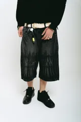 Bermuda de jean color negro con lavado acid wash gris. Presenta un corte baggy y detalles bordados en plateado y blanco que simulan ondas o llamas en la parte frontal superior e inferior.