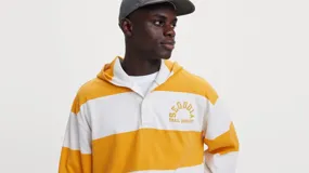 Sudadera Levi's de manga larga con diseño de rayas anchas horizontales blancas y amarillas, capucha integrada, cuello estilo polo con botones ocultos y bordado en el pecho. Corte holgado.