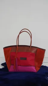 Bolso de fin de semana tipo tote, confeccionado en una combinación de lona y gamuza en tonos rojo, fucsia y marrón. Cuenta con doble asa de hombro, cierre superior y detalle de borlas colgantes en el lateral.