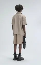 Camisa de manga corta color beige, estilo técnico, confeccionada en microfibra liviana. Cuenta con doble bolsillo frontal, uno con detalle de malla y otro con cierre, y broches en lugar de botones.