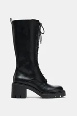 Bota de caña media con cordones frontales, confeccionada en material sintético negro. Presenta un tacón ancho en bloque y suela dentada con plataforma. Incluye cierre lateral y tirador trasero.