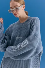 Sweater de tejido de punto grueso en color azul grisáceo, con escote redondo y diseño holgado. Presenta estampado gráfico en el frente y en una de las mangas, con terminaciones acanaladas en cuello, puños y ruedo.