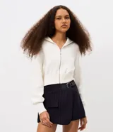 Campera cropped de tricot color blanco off-white, confeccionada en algodón con textura acanalada. Presenta cuello amplio con cierre metálico frontal, mangas largas con puños acanalados y cintura marcada con banda ancha.