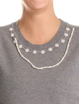 Buzo de tejido fino color blanco, con escote redondo decorado con un collar fijo de perlas y strass. Presenta mangas largas con un leve frunce en los hombros y puños acanalados.