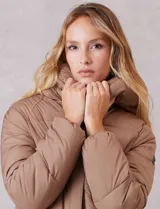 Campera puffer de corte recto y acabado mate, con diseño de bloques horizontales acolchados. Presenta capucha integrada con cordones de ajuste, cierre frontal metálico y bolsillos laterales invisibles. Incluye puños elásticos internos para mayor protección contra el frío.