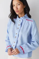 Camisa blanca de lino con cuello alto tipo gorguera, cierre frontal con botones de madera y detalles de ribetes en color mostaza en hombros y puños. Presenta volados en la botonera, cuello y puños.