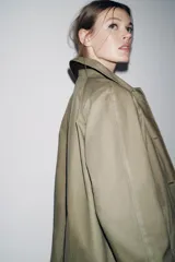 Trench coat de corte largo y diseño minimalista, confeccionado en tejido encerado color beige. Presenta cuello solapa, cierre frontal con botones oscuros y bolsillos laterales integrados.