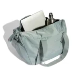 Cartera deportiva Adidas tipo tote, color verde claro, con doble asa de mano y correa ajustable para el hombro. Cuenta con cierre principal y bolsillo frontal con cierre, además del logo de la marca estampado en el frente.