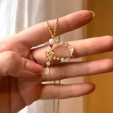 Collar dorado con dije en forma de letra P decorado con perlas, piedras blancas y rubíes. El dije tiene un dije colgante en forma de lágrima y una mariposa. La cadena es de eslabones pequeños.