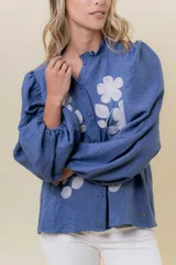 Camisa azul de calce relajado con bordado floral blanco en el frente. Presenta mangas largas amplias con ligero fruncido en el hombro y cierre frontal con botones.