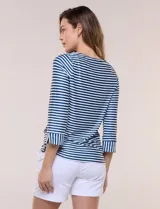 Remera de agua de silueta estilizada confeccionada en tejido técnico elastizado con protección solar UPF 50, con escote barco amplio y mangas 3/4 terminadas con puño en rayas verticales a contraste con pequeña abertura. Presenta aberturas laterales en el bajo decoradas con botones dorados grabados y ruedo recto de calce cómodo.