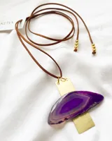 Collar con dije de piedra ágata morada y engarce de latón dorado, suspendido de un cordón de cuero marrón con cuentas doradas en los extremos.