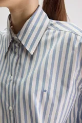 Camisa de calce relajado confeccionada en algodón, con diseño de rayas verticales en tonos beige y blanco. Presenta cuello clásico, cierre frontal con botones, puños abotonados y largo ligeramente cropped.