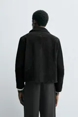 Cazadora de corte cropped fit, confeccionada en piel con acabado serraje (gamuza) de color negro. Presenta cuello solapa, manga larga, dos bolsillos de plastrón en la cadera y cierre frontal de botonadura.