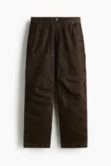 Pantalón estilo carpenter de corte holgado, confeccionado en tejido de algodón color marrón oscuro. Presenta tiro caído, bolsillos laterales de parche, trabilla funcional en una pierna y costuras reforzadas visibles sobre y debajo de las rodillas.