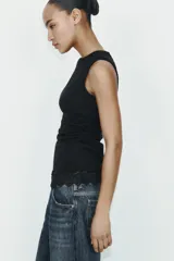 Camiseta ajustada (slim fit) color crema, con cuello redondo y mangas sisa. Presenta frunces laterales y un bajo asimétrico con detalle de encaje a tono.