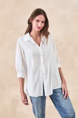 Camisa de corte holgado y fluido, color blanco, con cuello clásico, cierre frontal de botones y mangas tres cuartos con puños rectos.