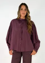 Camisa negra de corte holgado con cuello tipo bebé con volados y lazo de ajuste, cierre frontal con botones y mangas largas abullonadas con puños abotonados.