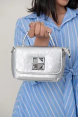 Cartera tipo baúl color plateado metalizado, con solapa y detalle de aplique piramidal metálico en el centro. Cuenta con asa corta de mano y correa para llevar al hombro.