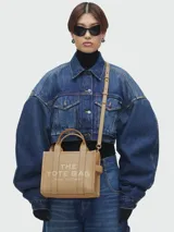 Cartera tipo tote de cuero granulado color tostado, con diseño estructurado y estampado frontal con la inscripción 'THE TOTE BAG MARC JACOBS'. Incluye doble asa superior y correa ajustable y extraíble para llevar al hombro o cruzada.
