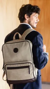 Mochila beige de tela canvas combinada con detalles en símil cuero marrón. Tiene apertura superior con cierre, bolsillo frontal con cierre, bolsillos laterales, bolsillos internos, asa corta de mano y correas acolchonadas regulables para la espalda.
