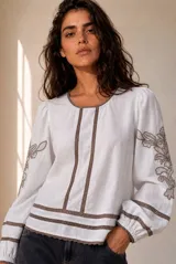Blusa blanca de manga larga con detalles de bordados florales en tono grisáceo y bandas decorativas en el frente y mangas. Cuenta con cuello redondo y puños elásticos.