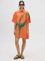 Camisa tipo vestido de corte oversize, confeccionada en viscosa. Presenta un diseño estampado con trazos abstractos en tonos verdes sobre una base naranja, cuello clásico, cierre frontal con botones y mangas cortas.