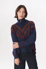 Buzo de tejido de punto en color azul marino, con cuello alto y cierre frontal parcial. Presenta un diseño con franjas en color marrón y detalles de líneas en rojo en el pecho, hombros y ruedo. Tiene corte holgado y mangas largas con puños acanalados.