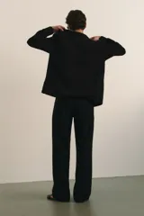 Pantalón negro de tejido suave con corte relajado, cintura elástica con cordón oculto y pliegues frontales. Presenta bolsillos laterales y bolsillos traseros ribeteados.