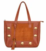 Bolso tipo tote de cuero vacuno color tabaco, con panel central de gamuza. Presenta detalles de tachas metálicas circulares doradas en los laterales y la base, y un colgante decorativo con borla en el frente. Cuenta con doble asa de mano y cierre superior.