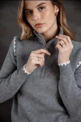 Buzo de punto acanalado color gris, con cuello alto y cierre frontal parcial. Presenta detalles de costuras decorativas con patrón de bloques en color blanco en los hombros, puños y ruedo.