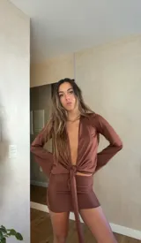 Vestido corto de satén color marrón, compuesto por una blusa de manga larga con escote profundo y nudo frontal, y una falda ajustada a juego.