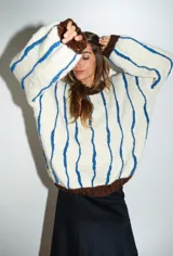 Buzo tejido a mano con lana merino color crudo, con cuello redondo y puños acanalados en marrón con lurex. Presenta bordados en relieve con rayas tipo cadenas en color azul.