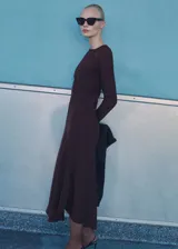 Vestido midi de color marrón oscuro, con cuello redondo, manga larga y diseño evasé. Presenta un detalle drapeado en la cintura y cierre de cremallera en la parte posterior.